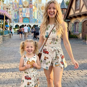 May include: Two people wearing matching cream-colored outfits with a Route 66 and car print. The outfits include a romper and a dress. The child is holding a Mickey Mouse-shaped pretzel. The background features a colorful building and people.