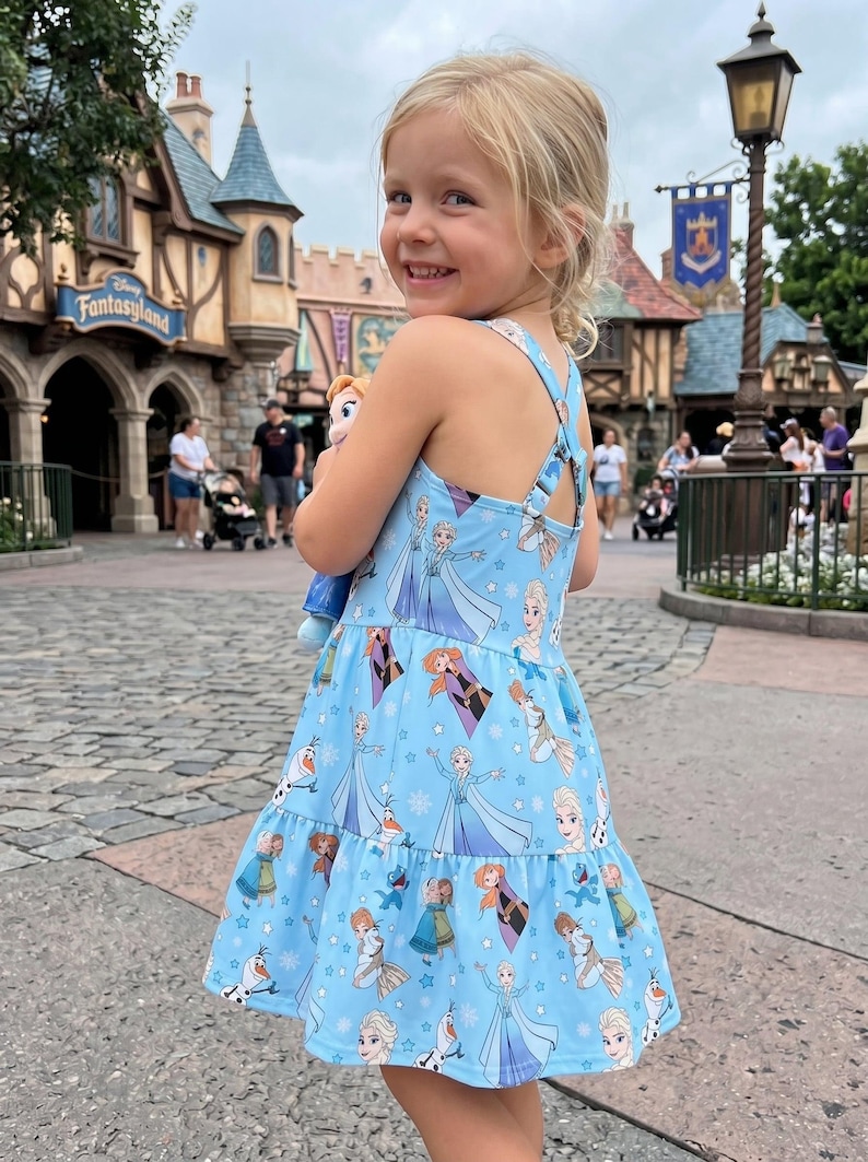 Pu&ograve; includere: Un vestito azzurro chiaro con personaggi del film Frozen. L'abito ha una gonna a balze e spalline incrociate. La bambina tiene in mano una bambola di Elsa.