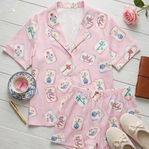 May include: Pink pajama set with a short-sleeved button-down shirt and matching shorts. The set is decorated with a repeating pattern of illustrated cards with floral and dragon motifs. A pair of cream-colored slippers with bows are also present.