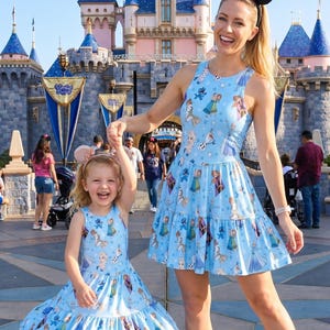 Pu&ograve; includere: Due donne con abiti azzurri abbinati con una gonna a balze e un motivo di personaggi di Frozen. Una donna indossa un cerchietto con le orecchie di Minnie Mouse. Sono in piedi davanti a un castello.
