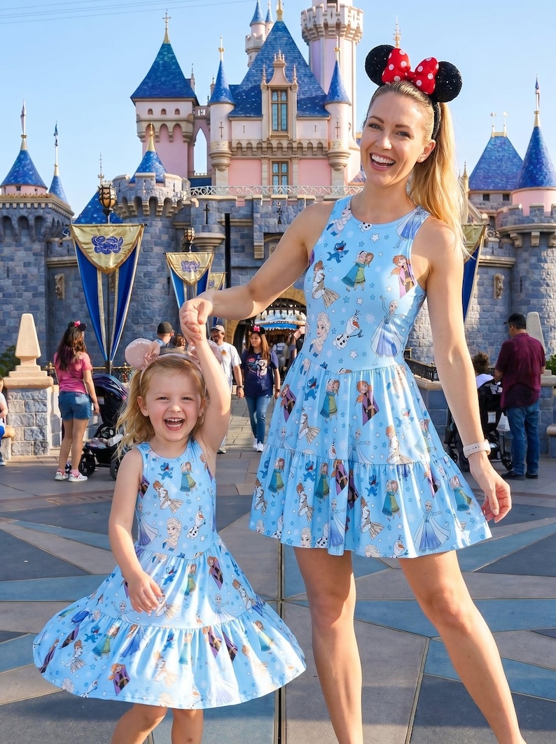 Pu&ograve; includere: Due donne con abiti azzurri abbinati con una gonna a balze e un motivo di personaggi di Frozen. Una donna indossa un cerchietto con le orecchie di Minnie Mouse. Sono in piedi davanti a un castello.