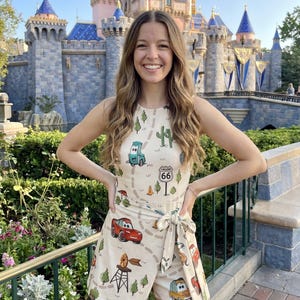 May include: A beige romper featuring a Cars-themed print with Route 66 signage. The sleeveless romper has a wrap-style design with a tie at the waist. The background includes a castle and lush greenery.