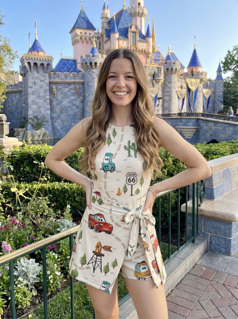 May include: A beige romper featuring a Cars-themed print with Route 66 signage. The sleeveless romper has a wrap-style design with a tie at the waist. The background includes a castle and lush greenery.