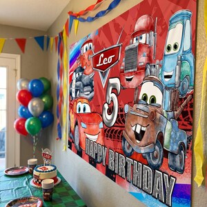 May include: A red banner with cartoon car characters and the text "Leo 5 HAPPY BIRTHDAY" is displayed. The scene includes a decorated table with a cake, plates, and balloons, suggesting a birthday party theme.