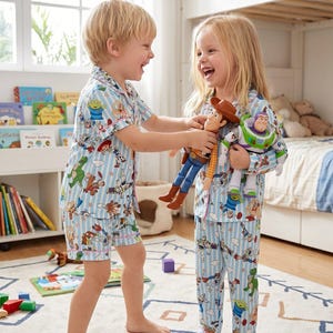 Puede incluir: Dos ni&ntilde;os con pijamas a rayas azules y blancas a juego con estampados de personajes de Toy Story. Un ni&ntilde;o sostiene mu&ntilde;ecos de Woody y Buzz Lightyear. Los pijamas incluyen una camisa de manga corta y pantalones cortos, y una camisa de manga larga y pantalones.