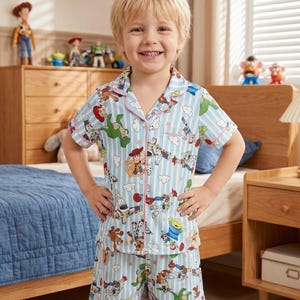 Puede incluir: Un ni&ntilde;o lleva un pijama azul claro con un estampado de Toy Story. La camisa de manga corta y los pantalones cortos presentan un patr&oacute;n de rayas verticales y ribetes rosas. El conjunto de pijama est&aacute; decorado con im&aacute;genes de personajes de la pel&iacute;cula Toy Story.
