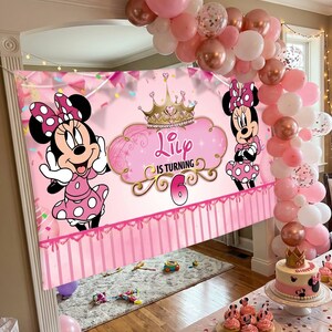 May include: A pink Minnie Mouse-themed birthday party scene. A banner with Minnie Mouse graphics and the text "Lily is turning 6" is displayed. Balloons in pink, white, and rose gold decorate the area, along with a cake and cupcakes.