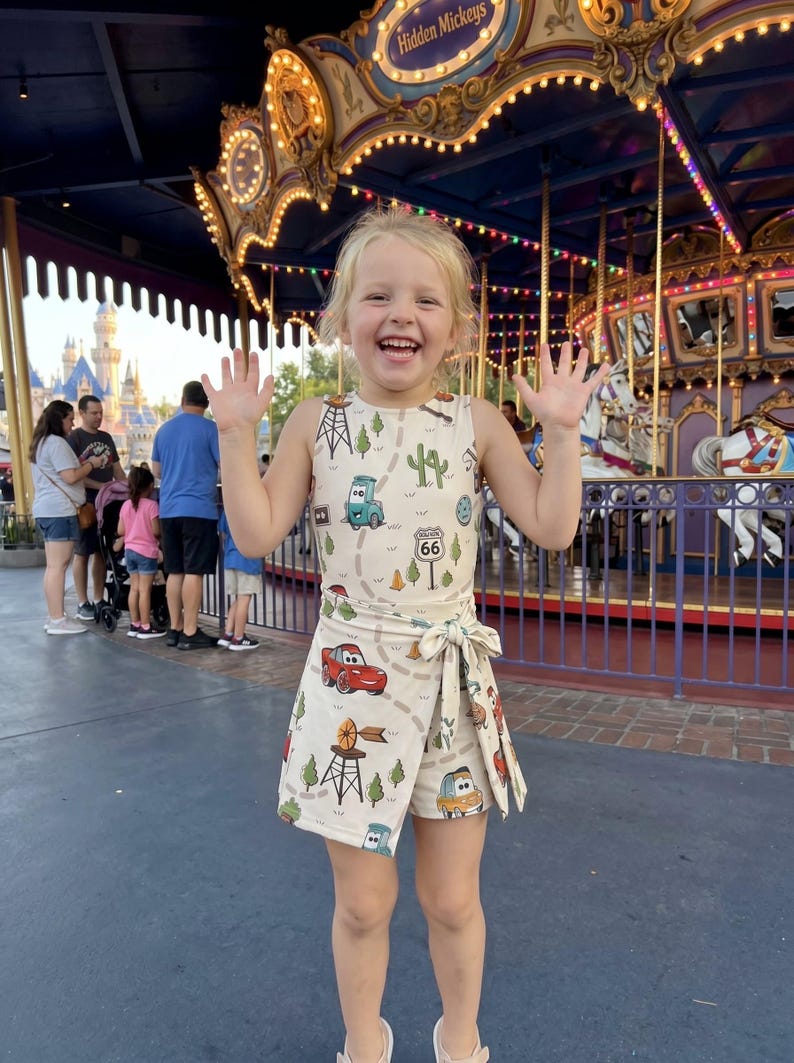 May include: A young child wearing a cream-colored romper with a Route 66 and car-themed print. The outfit includes a sleeveless top and wrap-around shorts. The child is smiling and waving in front of a carousel.