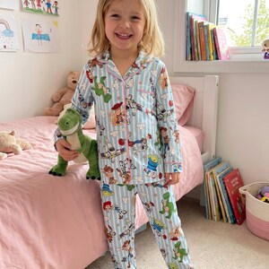Puede incluir: Conjunto de pijama azul claro para ni&ntilde;os con rayas blancas verticales y estampado de personajes de Toy Story. El conjunto incluye una camisa abotonada con ribete rosa y pantalones a juego. El ni&ntilde;o sostiene un peluche de dinosaurio verde.