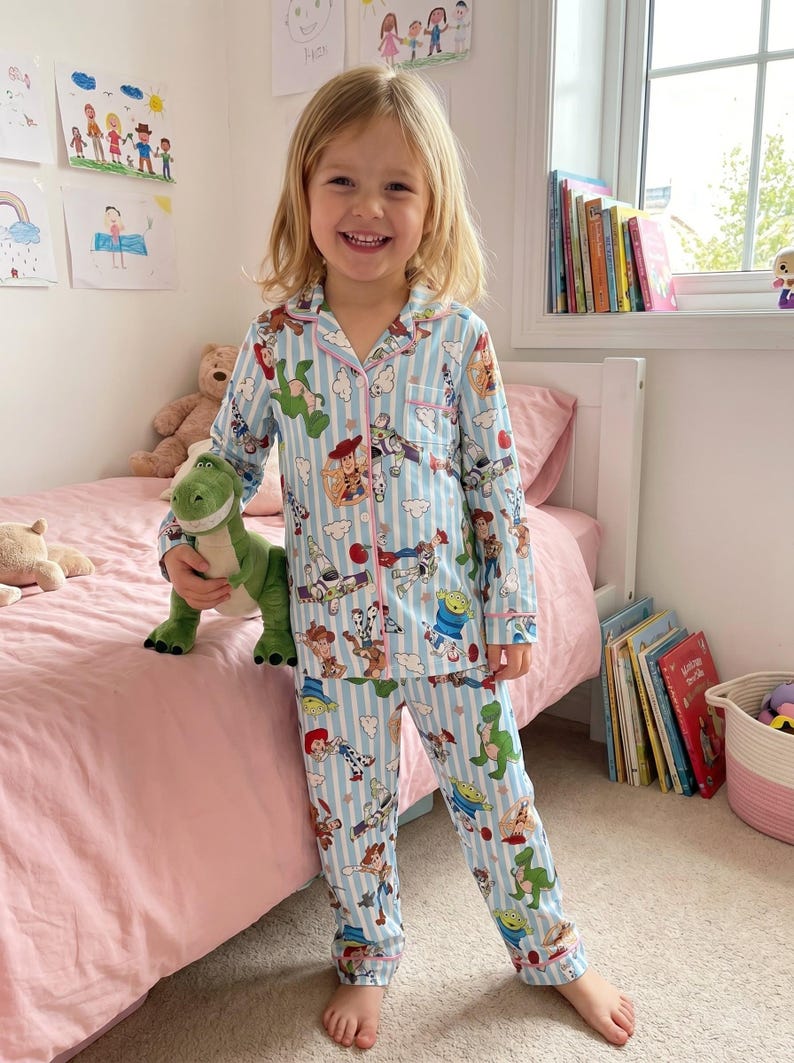 Puede incluir: Conjunto de pijama azul claro para ni&ntilde;os con rayas blancas verticales y estampado de personajes de Toy Story. El conjunto incluye una camisa abotonada con ribete rosa y pantalones a juego. El ni&ntilde;o sostiene un peluche de dinosaurio verde.