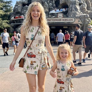 May include: Two women wearing matching beige rompers with a Route 66 and Cars-themed print. The rompers have a wrap-style skirt and are sleeveless. One woman carries a brown purse. The background shows a mountain and the sign "Matterhorn Bobsleds".