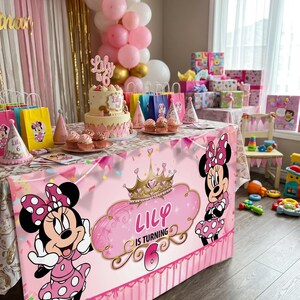 May include: A festive birthday party scene with a pink and gold color scheme. A table is decorated with a Minnie Mouse tablecloth, cake, cupcakes, and party favors. Balloons and gifts complete the celebratory atmosphere. The text on the tablecloth reads "Lily is turning 6."