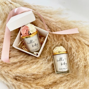 There are colorful artificial flowers on the glass candle made with soy wax. There is a customizable label on the cup. Next to the glass candle is a specially decorated box.