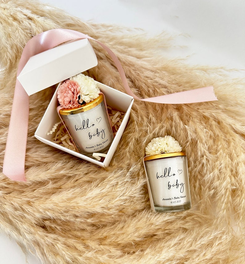 There are colorful artificial flowers on the glass candle made with soy wax. There is a customizable label on the cup. Next to the glass candle is a specially decorated box.