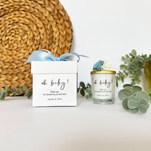 There are colorful artificial flowers on the glass candle made with soy wax. There is a customizable label on the cup. Next to the glass candle is a specially decorated box.