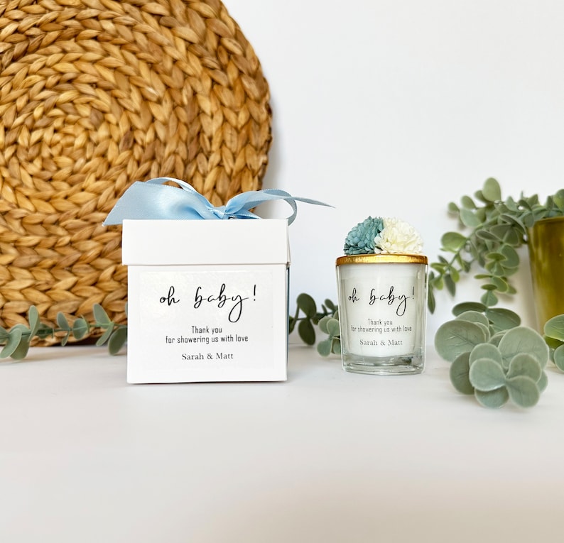 There are colorful artificial flowers on the glass candle made with soy wax. There is a customizable label on the cup. Next to the glass candle is a specially decorated box.