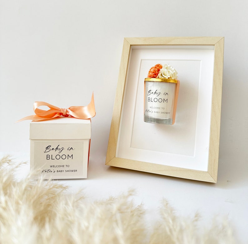 There are colorful artificial flowers on the glass candle made with soy wax. There is a customizable label on the cup. Next to the glass candle is a specially decorated box.