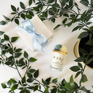 There are colorful artificial flowers on the glass candle made with soy wax. There is a customizable label on the cup. Next to the glass candle is a specially decorated box.