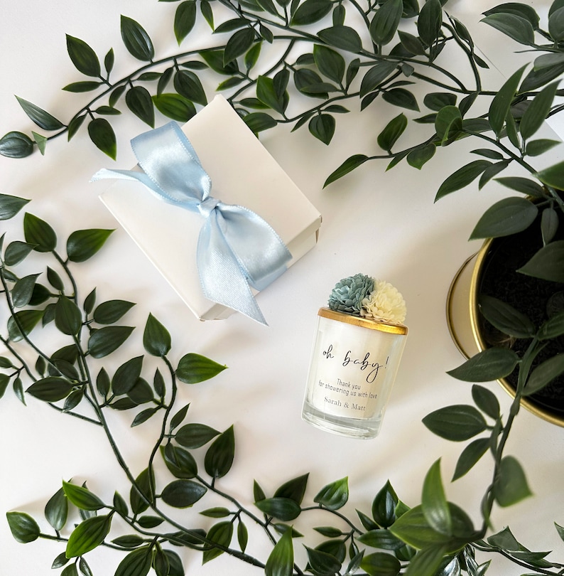 There are colorful artificial flowers on the glass candle made with soy wax. There is a customizable label on the cup. Next to the glass candle is a specially decorated box.