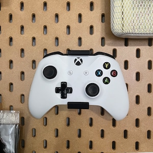 May include: A white Xbox controller with black buttons and a black grip. The controller is mounted on a pegboard wall.