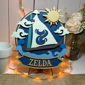 Peut inclure: Décoration en bois multicouche avec un bateau, un soleil et des nuages, avec le nom "ZELDA" en bas. Le design est principalement bleu, blanc et beige, avec un petit support en bois. Une tasse blanche et des fleurs sont en arrière-plan.