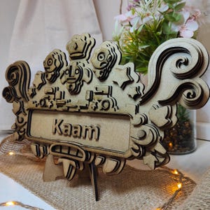 May include: A wooden sign with a layered design featuring a crown, stylized faces, and a nameplate that reads "Kaam". The sign is on a stand and has a whimsical, decorative style.