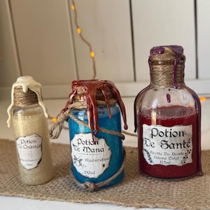 Può includere: Tre boccette di pozione in vetro con tappi di sughero e spago. La boccetta a sinistra contiene liquido dorato etichettato Potion De Chance 35ml. La boccetta centrale ha liquido blu etichettato Potion De Mana 100ml. La boccetta a destra ha liquido rosso etichettato Potion De Santé 125ml.