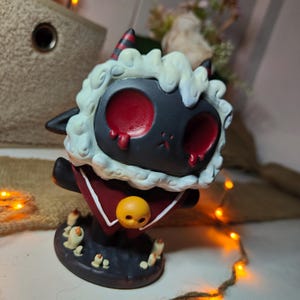 May include: A black and white lamb figurine with red eyes and horns. The lamb wears a red cape and a yellow skull pendant. The figurine sits on a black base with small candles.
