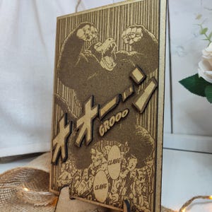 Puede incluir: Una pieza de arte de pared de madera con una ilustraci&oacute;n en blanco y negro de un gorila grande con el texto japon&eacute;s "オオゴリラ" y la palabra "GROOO" impresa debajo. La ilustraci&oacute;n est&aacute; rodeada de figuras m&aacute;s peque&ntilde;as y la palabra "CLANG" aparece dos veces en la imagen.