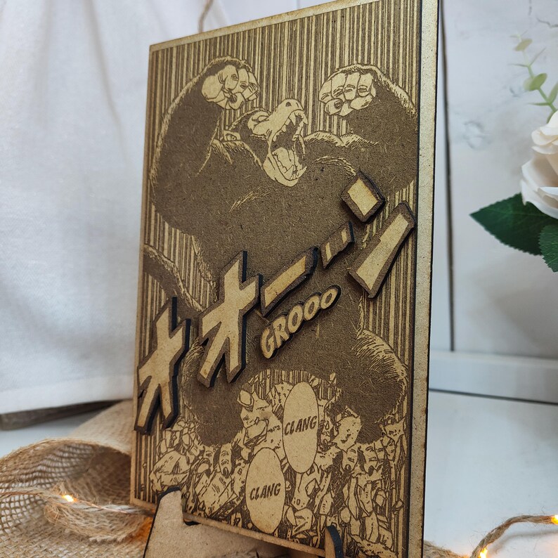 Puede incluir: Una pieza de arte de pared de madera con una ilustraci&oacute;n en blanco y negro de un gorila grande con el texto japon&eacute;s "オオゴリラ" y la palabra "GROOO" impresa debajo. La ilustraci&oacute;n est&aacute; rodeada de figuras m&aacute;s peque&ntilde;as y la palabra "CLANG" aparece dos veces en la imagen.