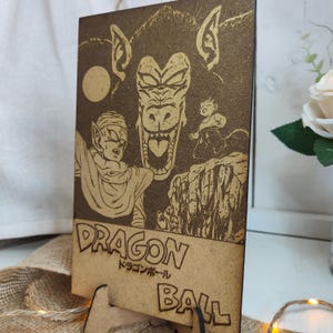 Puede incluir: Una placa de madera con una imagen grabada con l&aacute;ser de un personaje de la serie de anime Dragon Ball. La imagen muestra una gran criatura simiesca de aspecto feroz con una expresi&oacute;n amenazante. Un personaje m&aacute;s peque&ntilde;o est&aacute; de pie frente a la criatura, mirando hacia arriba con miedo. La placa es marr&oacute;n y tiene el texto "DRAGON BALL" y "ドラゴンボール" en caracteres japoneses.