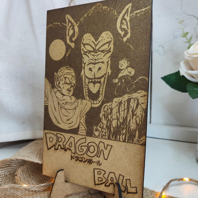 Puede incluir: Una placa de madera con una imagen grabada con l&aacute;ser de un personaje de la serie de anime Dragon Ball. La imagen muestra una gran criatura simiesca de aspecto feroz con una expresi&oacute;n amenazante. Un personaje m&aacute;s peque&ntilde;o est&aacute; de pie frente a la criatura, mirando hacia arriba con miedo. La placa es marr&oacute;n y tiene el texto "DRAGON BALL" y "ドラゴンボール" en caracteres japoneses.