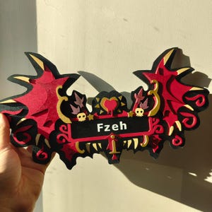 Puede incluir: Un letrero decorativo de varias capas en rojo, negro y dorado, con un diseño estilizado de alas de murciélago. El letrero incluye el nombre "Fzeh" en una placa rectangular negra, con motivos de calaveras y corazones.