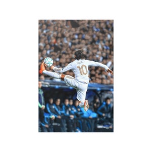 May include: A soccer player wearing a white jersey with gold trim and the number 10 on the back kicks a soccer ball in mid-air. The player is wearing white shorts and black socks. The player is surrounded by a crowd of people in the background.