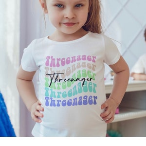May include: A white t-shirt with a rainbow design that says "Three-nager" in a retro font.