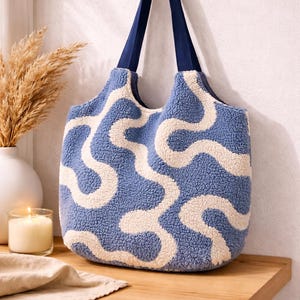 Könnte beinhalten: Blau-weiß gemusterte Tragetasche mit marineblauen Trägern. Die Tasche besteht aus einem weichen, strukturierten Material und hängt an einem Holzhaken. Die Tasche hat ein wellenförmiges Design.