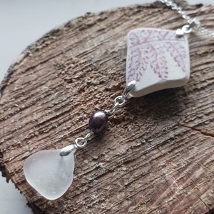 May include: A silver chain necklace with a white sea glass pendant, a dark brown pearl, and a white sea glass pendant with a pink floral design.