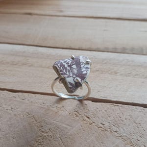 Pode incluir: Anel de prata com uma pedra triangular com um desenho floral bordô sobre fundo creme. A montagem é ajustável e a pedra é presa por garras de prata. Uma joia única.