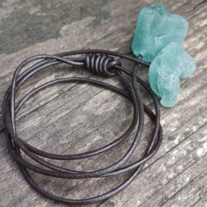 May include: A brown leather cord necklace with two teal sea glass pendants. The necklace is tied in a knot and the sea glass is shaped like hearts.