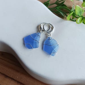 Puede incluir: Un par de pendientes con piezas de vidrio marino azul esmerilado y liso, envueltas en un fino alambre de tono plateado. Cada pieza de vidrio está suspendida de un pequeño aro circular de tono plateado, creando un diseño de joyería sencillo.
