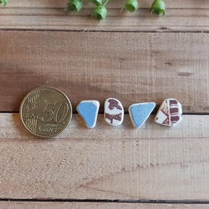 Op de afbeelding: Vier kleine keramische tegels met een witte achtergrond en blauwe en bruine accenten. De tegels zijn in een rij op een houten oppervlak gerangschikt.