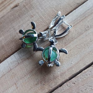 May include: A pair of silver turtle earrings with green glass beads inside. The earrings have a hoop closure.