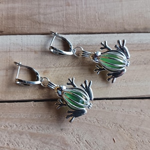Peut inclure: Une paire de boucles d'oreilles en forme de grenouille argentées avec des yeux en verre vert. Les grenouilles pendent à des créoles argentées.