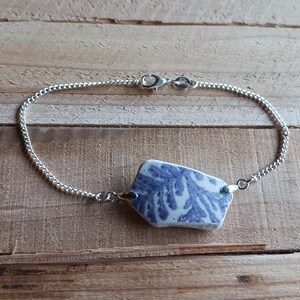 Puede incluir: Un brazalete de cadena de plata con un colgante de cerámica azul y blanco. El colgante tiene una forma irregular y única, con un patrón de remolino.