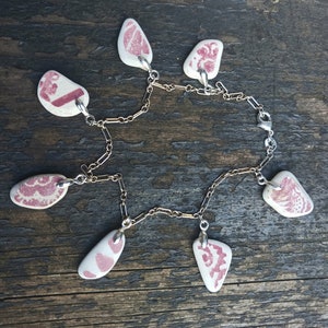 Puede incluir: Un collar de cadena de plata con seis colgantes. Cada colgante es una piedra blanca con un diseño rosa. Los colgantes están conectados a la cadena con anillos de plata.