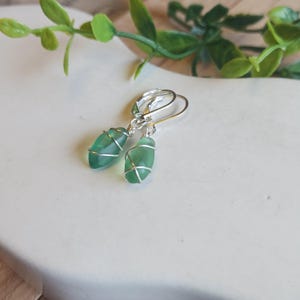 May include: A pair of drop earrings featuring smooth, frosted green sea glass pieces wrapped in thin silver-toned wire. The earrings are attached to silver-toned lever-back hooks, creating a simple and clean design for these dangling accessories.