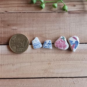 May include: A collection of small, irregularly shaped ceramic pieces with blue, pink, and white floral and geometric patterns. A 50 Euro cent coin is next to the pieces for size comparison. The items are arranged on a wooden surface.