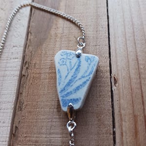 Puede incluir: Un collar de cadena de plata con un colgante de cerámica azul y blanco con un diseño floral. El colgante tiene forma de lágrima y tiene un cierre de plata.
