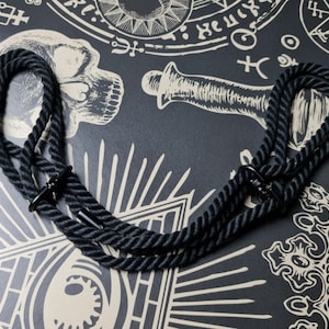 Könnte beinhalten: Eine schwarze Seilkette mit Metallbeschlägen. Die Kette besteht aus dickem, gedrehtem Seil und ist herzförmig angeordnet. Der Hintergrund zeigt eine dunkle Oberfläche mit weißen okkulten Symbolen.