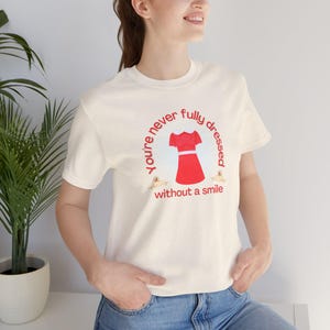 Puede incluir: Una camiseta blanca con un gráfico rojo que dice "You're never fully dressed without a smile". El gráfico incluye un vestido rojo con cuello blanco y cinturón blanco, y dos pequeños perros amarillos a cada lado del vestido.